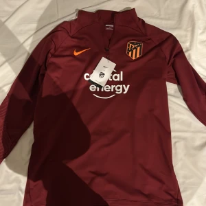 Atletico Madrid långärmad Nike tröja - Slim fit fotbollströja från Nike i mörkröd färg med Atletico Madrid-logga på bröstet och orange swoosh. Tröjan har lång ärm med subtilt mönster på underarmarna och vit tryckt sponsortext. Materialet är lätt och andas, perfekt för träning.