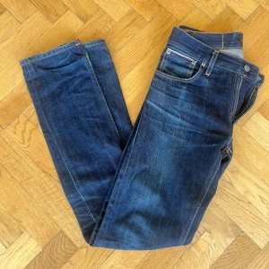 Dry selvage jeans från Nudie - Mörkblå dry selvage jeans från Nudie. Jeansen har fått en snygg tvätt efter att jag använt dem vilket ger dem personlighet men har tyvärr växt ur dem nu och hoppas att någon annan kan ge dem nytt liv. Storlek W28.