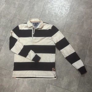 Svartvit randig pikétröja Gant - Säljer en klassisk pikétröja från Tommy Hilfiger med breda svarta och vita ränder. Tröjan har lång ärm, krage och knappslå framtill. Detalj med liten patch längst ner. Perfekt för dig som gillar sportig och stilren look. Vintage 