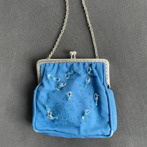 Blå kuvertväska med pärldetaljer - Söt blå kuvertväska med silverfärgad kedja och ram. Väskan är dekorerad med små blå pärlor och genomskinliga pärldroppar på framsidan. Materialet är mjukt och har en lyxig känsla, perfekt för att lyfta din outfit. Insidan är fodrad i ljusblått tyg.