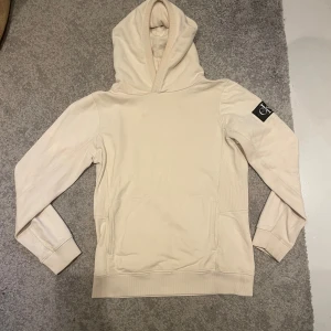 Beige hoodie från Calvin Klein Jeans - Snygg beige hoodie från Calvin Klein Jeans med stor CK-logga på ena ärmen. Klassisk modell med huva och känguruficka framtill. Mjuk bomullskvalitet och ribbade muddar vid ärmslut och nederkant. Perfekt för en chill och stilren look.