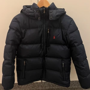 Marinblå pufferjacka från Polo Ralph Lauren - Marinblå pufferjacka från Polo Ralph Lauren med huva, dragkedja och två sidofickor med zip. Jackan har det klassiska röda Polo-logot broderat på bröstet och quiltad design. Perfekt för kalla dagar och riktigt snygg streetstil. Jackan är i väldigt fint skick.
