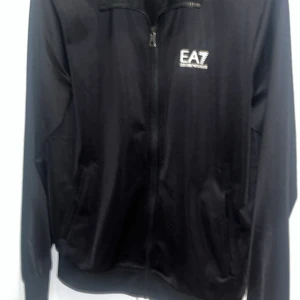 Svart vindjacka från EA7 Emporio Armani - Svart vindjacka från EA7 Emporio Armani med stilren design och logga på bröstet. Jackan har dragkedja framtill, två sidofickor och ribbade muddar. Materialet är lätt och syntetiskt, perfekt för en sportig look. Långärmad modell med diskreta detaljer på ryggen. XS passar S. Jätte bra skick
