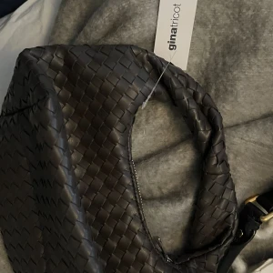 Brun flätad axelväska från Gina Tricot - Snygg brun axelväska från Gina Tricot med flätad design och mjuk form. Väskan har dragkedja upptill och ett kort handtag som gör den enkel att bära över axeln. Perfekt för dig som gillar trendiga detaljer och stilren look.