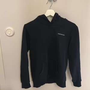 Mörkblå hoodie Peak Performance 160 - Mörkblå hoodie från Peak Performance i storlek 160. Klassisk design med huva, magficka och diskret logotyp på bröstet. Mjuk och skön känsla, perfekt för chill eller träning. Långärmad och med ribbade muddar vid ärmslut och nederkant.