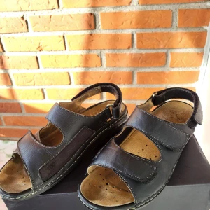 Svarta sandaler från Scholl i skinn - Svarta sandaler från Scholl med breda remmar i äkta skinn och justerbar kardborreknäppning. Sval design med öppen tå och perforerad innersula för extra komfort. Snygga kontrastsömmar och stabil platt sula, perfekta för varma dagar.