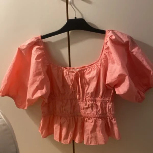 Rosa blus med puffärmar, strl S från Hm  - Säljer en söt rosa blus i storlek S med puffiga korta ärmar och volang nedtill. Toppen har en rundad halsringning med knyt framtill och smockad midja som ger en snygg siluett. Perfekt för dig som gillar romantisk och trendig stil.