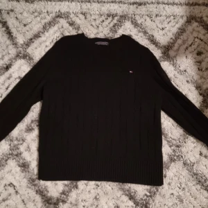 Svart stickad tröja Tommy Hilfiger - Svart stickad tröja från Tommy Hilfiger i storlek S. Tröjan har kabelstickat mönster, rund hals och långärmade ärmar. Liten Hilfiger-logga broderad på bröstet. Perfekt för dig som gillar klassisk och enkel stil.