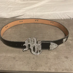 Svart skärp från Tuttolente - Säljer ett svart skärp från Tuttolente med bred form och silverdetaljer. Bältet har en stor, blingad spänne med bokstäver och en dekorerad örn i silver. Hela bältet har en lyxig vibe. Använt 1 gång