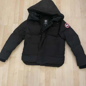 Svart dunjacka från Canada Goose - Säljer en svart dunjacka från Canada Goose med huva och klassisk logga på ärmen. Jackan har dragkedja och stora fickor framtill, perfekt för kalla vinterdagar. Stilen är clean och jackan är riktigt varm och skön.