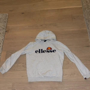 Grå hoodie från Ellesse med logga - Snygg ljusgrå hoodie från Ellesse med stor logga framtill i svart, orange och rött. Tröjan har huva, magficka och ribbade muddar. Riktigt fräsch, pris går att diskuteras :)