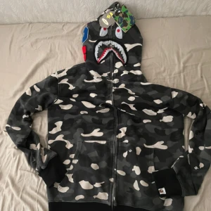 BAPE Shark Camo Hoodie svart/grå - Säljer en ikonisk BAPE Shark Hoodie i svart och grå camo med dragkedja hela vägen upp i huvan. Tröjan har broderade detaljer med hajansikte och WGM-bokstäver i rött, blått och orange. Klassisk streetwear med cool camomönster och feta detaljer.