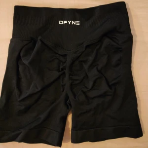 Svarta träningsshorts DFYNE - Svarta träningsshorts från DFYNE med ribbad midja och tight passform. Perfekta för gymmet eller löpning, tillverkade i mjukt och stretchigt syntetmaterial som sitter skönt mot kroppen. Snygg logga framtill i vitt.