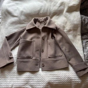 kappa från Kappahl - Kort beige brun jacka från Kappahl med stora fickor framtill, klassisk krage och knäppning med knappar. Snygg boxig passform och stilrena detaljer vid ärmslut. Perfekt för lager-på-lager under höst och vinter.