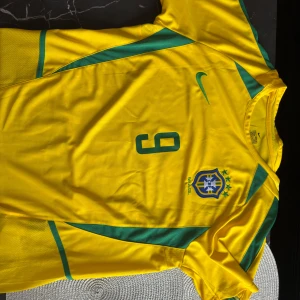 Brasilien Ronaldo 9 Nike fotbollströja - Säljer en ikonisk Brasilien fotbollströja från Nike i storlek L med Ronaldo och nummer 9 på ryggen. Tröjan är gul med gröna detaljer, har korta ärmar och är tillverkad i lätt, ventilerande polyester. Perfekt för dig som älskar klassiska landslagströjor och legendariska spelare. priset är ej hugget i sten