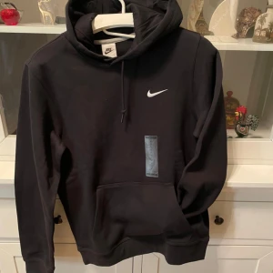 Svart hoodie från Nike med logga - Svart hoodie från Nike med klassisk vit swoosh-logga på bröstet. Tröjan har huva med snörning och en stor magficka framtill. Perfekt för en chill och sportig stil. Tillverkad i mjuk bomullsmix för extra komfort. Pris kan diskuteras