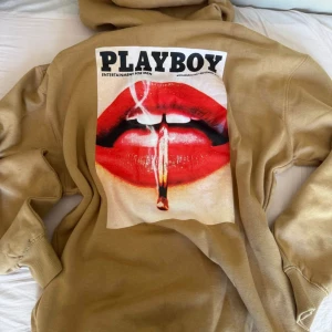 Beige Playboy hoodie med tryck - Snygg oversized beige hoodie från Playboy x Missguided med stor grafisk print på ryggen av röda läppar och tändsticka. Klassisk huva, känguruficka och PLAYBOY-logga fram. Perfekt för dig som vill sticka ut med streetstyle-vibe. 