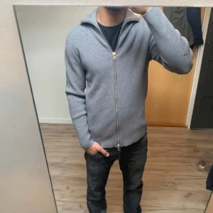 Grå kofta från Ljung by Marcus Larsson - Säljer en stilren grå ribbad kofta från Ljung by Marcus Larsson. Koftan har hel dragkedja framtill, hög krage och är långärmad. Perfekt för lager-på-lager och passar till både jeans och chinos. Materialet är mjukt och stickat för extra komfort. Nypris:1400kr Mitt pris:449Kr