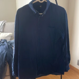 Mörkblå manchesterskjorta från Uniqlo - Säljer en stilren mörkblå manchesterskjorta från Uniqlo i storlek L.  Känns som M i storlek. Skjortan har klassisk krage, bröstficka och knäppning framtill. Perfekt för dig som gillar en enkel men snygg look. Materialet är mjukt och ger en skön känsla.