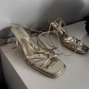 Guldiga klackskor med snörning Boohoo - Snygga klackskor från Boohoo i metallic guld med fyrkantig tå och smala remmar som snöras runt benet. Skorna har låg klack och ett ormskinnsmönster som ger extra edge. Perfekta för dig som vill sticka ut med en trendig look.