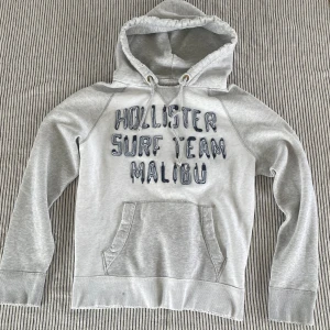 Grå hoodie från Hollister med tryck - Snygg ljusgrå hoodie från Hollister med broderat ’Hollister Surf Team Malibu’ på bröstet och en stor siffra 2 på ryggen | Size: M | Defekter förekommer men ska se vintage ut | Väldigt sällsynt och svår att få tag på!