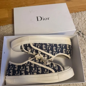 Dior sneakers i blått och vitt - Snygga Dior sneakers med blått och vitt monogrammönster, vita snören och detaljer. Ovandel i textil med tydligt Dior-logga, vit platt sula och unik stjärndetalj undertill. Perfekta för dig som vill sticka ut med lyxig streetstyle. Boxen medföljer i köpet. Köpt i andrahand och har därav inget äkthetsbevis.