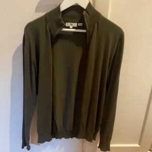 Olivgrön kofta från Mo - Snygg olivgrön kofta med dragkedja från Mo i mjukt material. Enkel och stilren design, med ribbad krage och muddar. Perfekt att slänga över en t-shirt för en chill look. Långärmad och passar till många outfits.