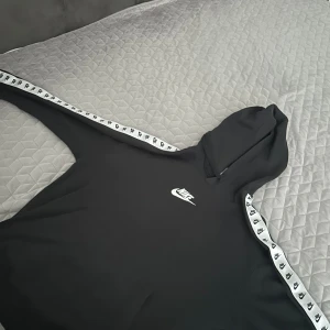 Svart Nike hoodie med logoband - Svart hoodie från Nike med vit broderad logga på bröstet och vita band med svarta Nike-loggor längs ärmarna. Klassisk huva och känguruficka framtill. Perfekt för en sportig och avslappnad stil.