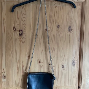 Svart axelväska med kedja och nitar - Snygg svart axelväska i skinn med silverfärgad kedjerem och coola nitar längs kanterna. Väskan har dragkedja upptill och är perfekt för att bära det viktigaste. Enkel men edgy design som lyfter vilken outfit som helst.