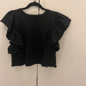 Svart topp med volangärmar - Säljer en svart topp med breda volangärmar och rund halsringning. Modellen är croppad och har en loose passform som ger en avslappnad vibe. Perfekt att styla med höga jeans eller kjol för en cool look.