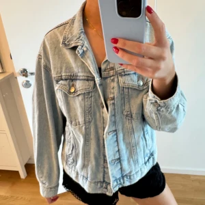 Ljusblå jeansjacka - Säljer en ljusblå jeansjacka med klassisk krage och knappar framtill. Jackan har två bröstfickor med lock och guldiga knappar, samt långa ärmar. Perfekt för en chill och avslappnad stil.