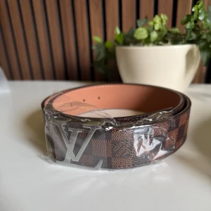 Louis Vuitton rutigt brunt bälte (3) - Snyggt och brett bälte från Louis Vuitton med klassiskt rutigt mönster i mörkbrunt och brunt. Bältet har en stor LV-logga i silverfärgad metall som spänne och är gjort i exklusivt skinn. Perfekt accessoar för att lyfta din outfit.
