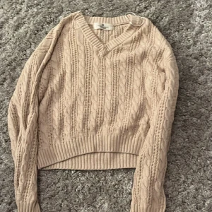 Beige kabelstickad tröja H&M - Säljer en beige kabelstickad tröja från H&M med v-ringning och ribbade muddar. Tröjan har lång ärm och klassiskt stickat mönster som ger en chill och snygg vibe. Perfekt att slänga på sig när det är lite kyligt ute. Perfekt för hösten!