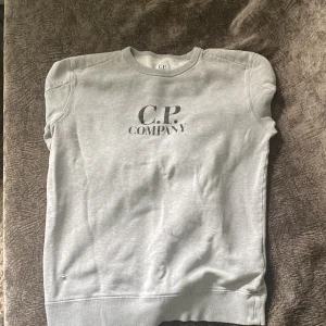 Grå sweatshirt från C.P. Company - Grå sweatshirt från C.P. Company med klassisk logga på bröstet och den ikoniska svarta linsen på ärmen. Tröjan har rund halsringning och är tillverkad i mjuk bomull. Tröjan har en litet hål vid magen men syns knappast, pris kan diskuteras. Hör av dig vid frågor!