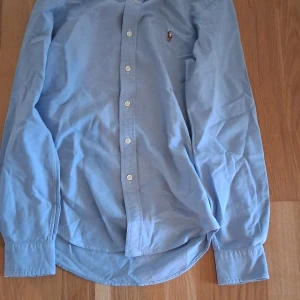 Ljusblå skjorta från Polo Ralph Lauren - Klassisk ljusblå skjorta från Polo Ralph Lauren med button down-krage och broderad logga på bröstet. Skjortan har långa ärmar, vita knappar och är tillverkad i mjuk bomull. Perfekt för en stilren och avslappnad look. 