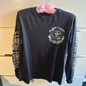 Sons of Anarchy långärmad tröja - Svart långärmad tröja med stort Sons of Anarchy-tryck på ryggen och mindre logga fram. Texten 'SAMCRO FOREVER' på ena ärmen. Skön passform och coolt tryck för dig som gillar serien. Perfekt till jeans eller svarta byxor.