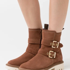 Cognac mockaboots från Tamaris - Snygga cognacsfärgade boots från Tamaris i mocka med två guldfärgade spännen och grov sula. Dragkedja på insidan för enkel påtagning. Perfekta för dig som gillar en cool och stilren look till höst och vinter. Står 39, men det är 40 i verkligheten. 