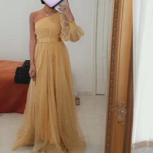 Gul långklänning med bälte och slits - Säljer en maxad gul långklänning med glittrigt prickigt tyg, bred bälte i midjan och en dramatisk oneshoulder-ärm med volym. Klänningen har en snygg slits framtill och markerad midja. Perfekt för dig som vill sticka ut med en glamorös look. Från Mr.& Mrs.