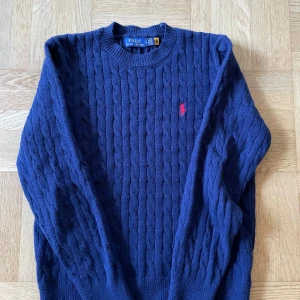 Polo Ralph Lauren tröja - Kabelstickad marinblå tröja från Polo Ralph Lauren med klassisk rund hals och broderad röd logga på bröstet. Långärmad och ribbade muddar för en snygg passform. Perfekt för dig som gillar stilren och tidlös design.