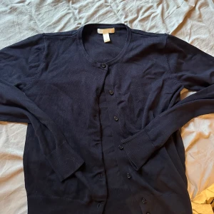 Mörkblå kofta från H&M Basics XS - En klassisk mörkblå kofta från H&M Basics i storlek XS. Koftan har rund halsringning, långa ärmar och knäppning framtill med svarta knappar. Perfekt att slänga över en t-shirt eller linne för en enkel och stilren look.