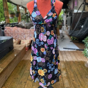 Blommig långklänning från MNG Suit S - Svart klänning med färgglada blommor. Klänningen har en v-ringning och smala axelband som korsas i ryggen. Den har en åtsittande midja och en utsvängd kjol som slutar strax under knät. Underkjol. ﻿Knytband i midja ﻿Bortklippta tvättråd ﻿Polyester ﻿Längd: 107 cm ﻿Bystvidd: 35 cm  Inga bilder när den sitter på mig! Katter finns i hemmet! 01,S