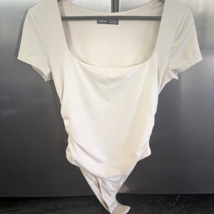 Vit kortärmad bodysuit från SHEIN - Säljer en stilren vit bodysuit från SHEIN med korta ärmar och fyrkantig urringning. Toppen är figurnära och har en clean look som passar perfekt till jeans eller kjol. Gjord i mjuk bomullsmix för en bekväm känsla hela dagen.