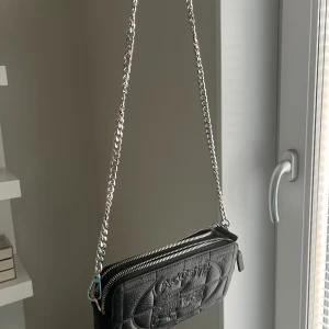 Svart axelväska från Lacoste i skinn - Säljer en svart axelväska från Lacoste i äkta skinn med silverfärgad kedjerem. Väskan har quiltad struktur, präglad logga framtill och dubbla dragkedjor upptill. Perfekt storlek för det viktigaste och ger en clean vibe till din outfit.