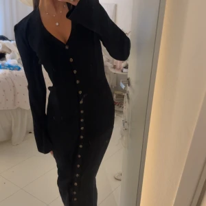 Svart långklänning med knappar - Snygg svart långklänning med v-ringning och långa ärmar. Klänningen har en rad dekorativa knappar framtill och är figurnära med lätt utsvängda ärmslut. Materialet ser ut att vara ribbad och stretchigt, vilket ger en cool och modern vibe.