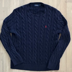 Mörkblå kabelstickad tröja Polo Ralph Lauren - Kabelstickad mörkblå tröja från Polo Ralph Lauren med klassisk röd broderad logga på bröstet. Tröjan har rund halsringning, långa ärmar och ribbade muddar. Perfekt för dig som gillar preppy stil och vill ha något både snyggt och bekvämt.