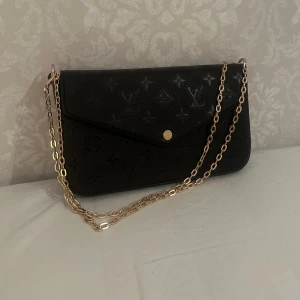 svart axelväska i skinn - Säljer en svart axelväska med klassiskt monogrammönster i präglat skinn. Väskan har guldfärgad kedja och knäppning framtill. Insidan är röd med plats för kort och småsaker. Perfekt för dig som vill ha något stilrent och lyxigt.