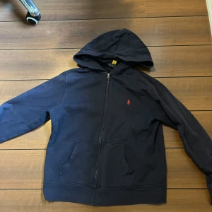 Marinblå hoodie från Polo Ralph Lauren - Säljer en marinblå hoodie från Polo Ralph Lauren med dragkedja och huva. Tröjan har en klassisk röd broderad logga på bröstet, känguruficka och är tillverkad i mjuk bomull. Perfekt för chill dagar eller när du vill ha en clean och stilren look. Storlek L barn passar dig som är 160-165. Säljer eftersom den är för liten. Hör gärna av dig vid prisdiskusion eller andra frågor😀
