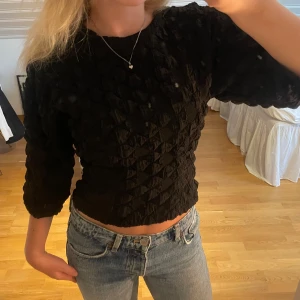 Svart strukturerad blus med trekvartsärm - Säljer en svart blus med coolt strukturerat tyg och trekvartsärmar. Toppen är croppad och har en liten öppning i ryggen. Perfekt för dig som vill ha något unikt och trendigt i garderoben.