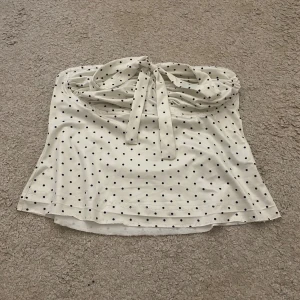 Vit prickig bandeau tubtopp med knyt - Supersöt vit bandeau tubtopp med svarta prickar och knyt fram. Toppen är ärmlös och har en lätt utsvängd passform. Perfekt för varma dagar och ger en retrovibe. Mjuk och stretchig känsla, troligen i bomull eller polyesterblandning. Jag har sprättat upp toppen längst ner då den ska bli lite längre.