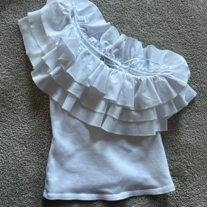 Vit topp från zara - Supersöt vit oneshoulder topp från Zara med stora volanger längs halsringningen. Toppen är ärmlös och har en snygg asymmetrisk design som verkligen sticker ut. Perfekt för dig som vill ha något unikt och trendigt i garderoben.